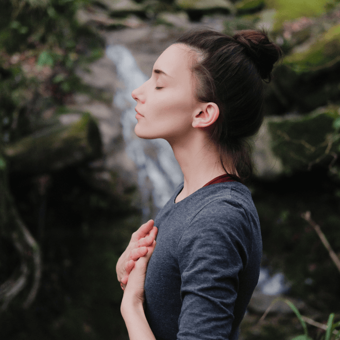 Matt Fraser, wearing a gray shirt, stands outdoors with his eyes closed and hands on his chest. He sports a bun hairstyle, and the background is a blurred natural setting featuring rocks and greenery.