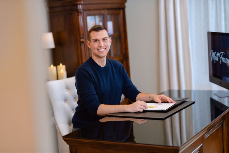 Matt Fraser, in a dark sweater, sits at a desk with an open notebook and pencil, smiling at the camera. A monitor and candles are in the background.