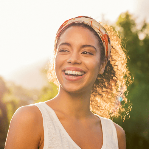 Woman with curly hair and headband, smiles brightly outdoors with sunlight and greenery in the background.