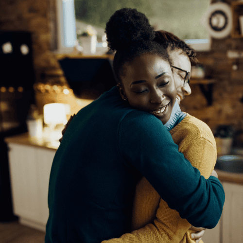 Two people hugging in a warmly lit kitchen, both smiling and wearing cozy sweaters.