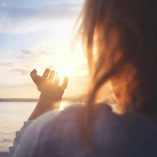 A woman extends her hand toward the sun at sunset by the water, creating a silhouette effect with warm light and a soft-focus background.