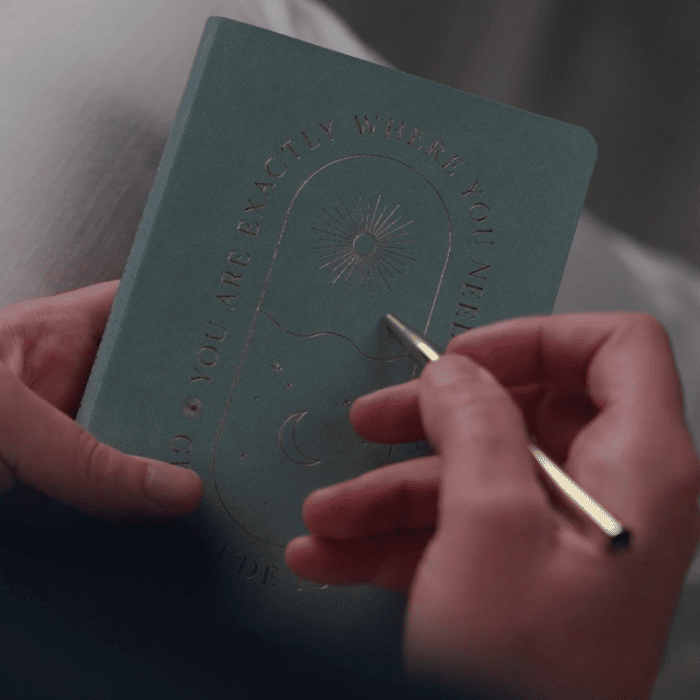 Matt Fraser is holding a teal notebook with an embossed celestial design and text. He is using a gold pen while sitting in a bright setting.