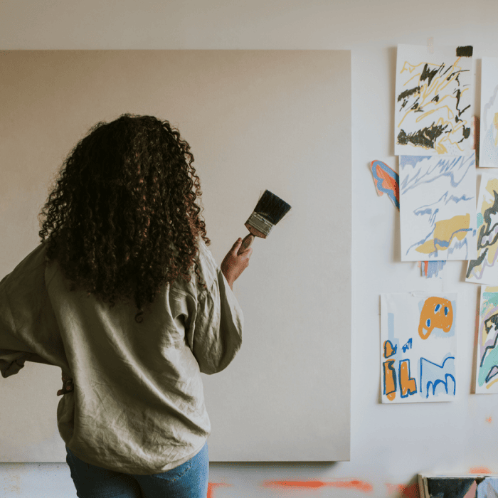Matt Fraser, with curly hair, is holding a brush and facing a large blank canvas. Colorful artworks are displayed on the adjacent wall.