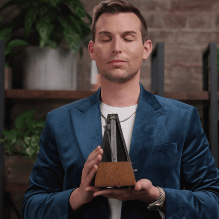 Matt Fraser, wearing a blue blazer, stands with his eyes closed, holding a metronome. The background features plants and shelves against a brick wall.