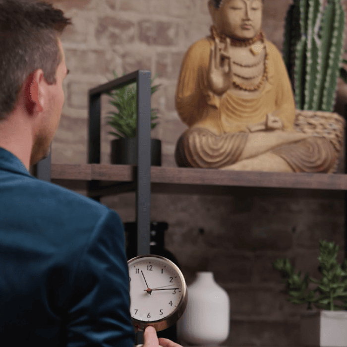 Matt Fraser is holding a clock while facing shelves with a Buddha statue, plants, and decorative items.