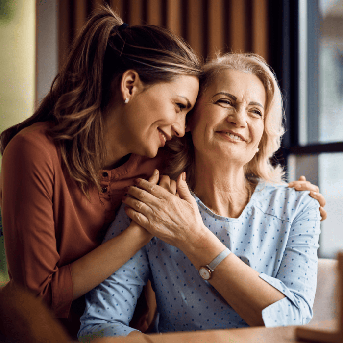 A young woman embraces an older woman with a smile indoors. Both appear happy and content.