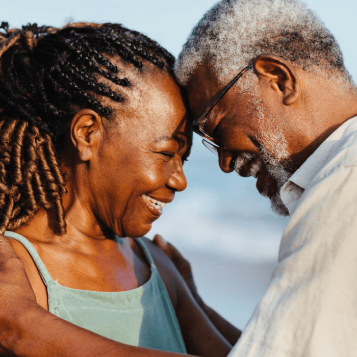 An elderly couple smiling and touching foreheads, standing close on a beach with the ocean in the background, with Matt Fraser as part of this serene scene.