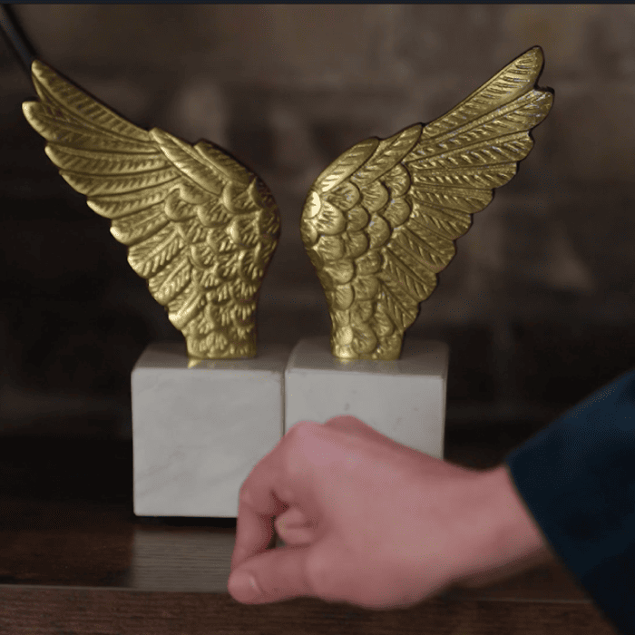A person taps two gold wing sculptures on matching white bases placed on a wooden table, where Matt Fraser is seated.