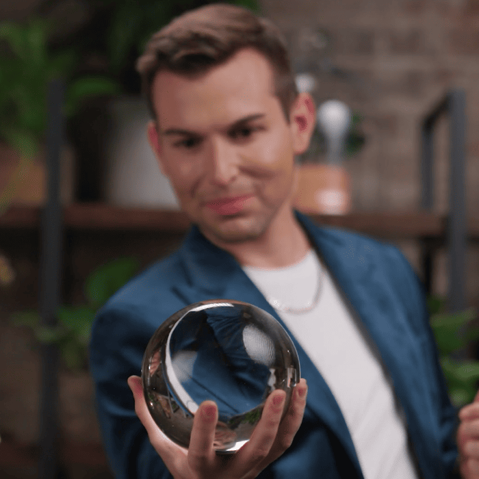 Matt Fraser, in a blue blazer, is holding a clear glass orb, with a blurred background of shelves and plants.
