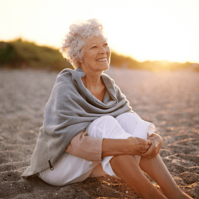 Elderly woman with curly white hair sits on a beach at sunset, wearing a gray shawl and light clothing, gazing into the distance with a gentle smile.