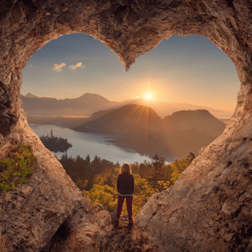 A woman is standing inside a heart-shaped cave opening, overlooking a scenic view of mountains, a lake, and a sunrise.
