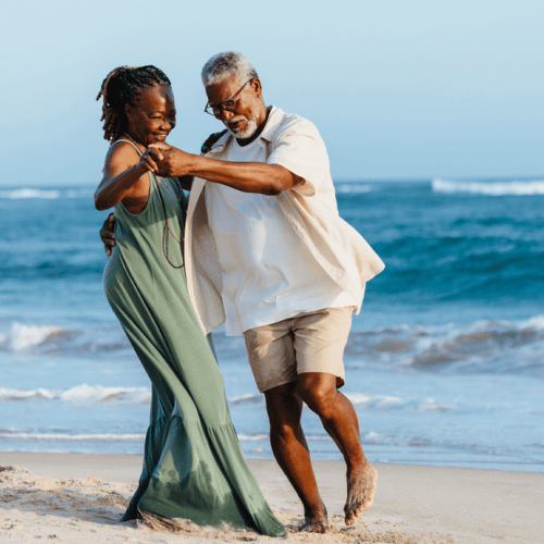 An older couple dances joyfully on a sandy beach by the ocean, with Matt Fraser wearing a light shirt and shorts, and the woman in a long green dress. Waves and blue sky are in the background.