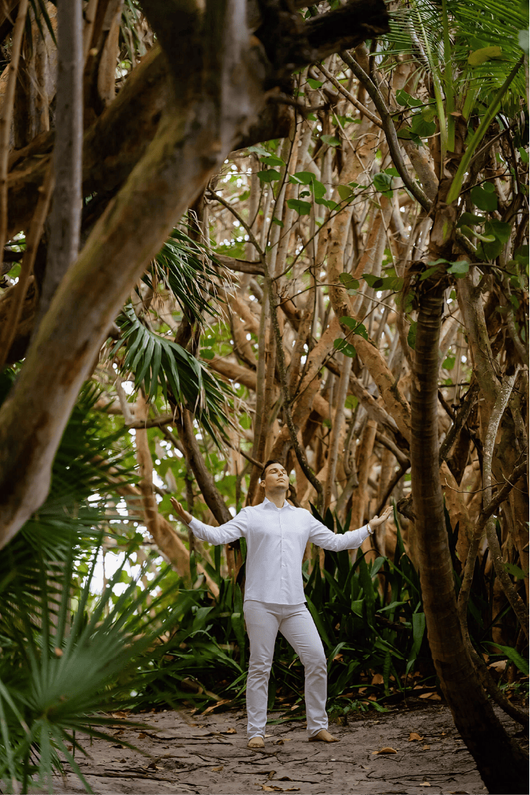 Matt Fraser, dressed in white clothing, stands with his arms outstretched in a dense, tropical forest setting.