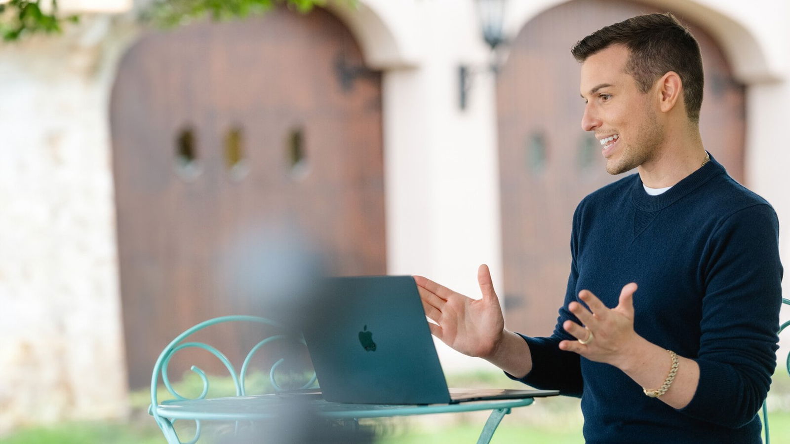 Matt Fraser sits outside at a table, engaged in a video call on a laptop, gesturing with his hands while speaking.