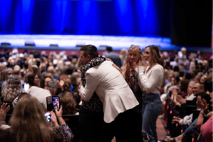 Matt Fraser, wearing a white jacket, hugs someone in a crowded auditorium as people around them clap.