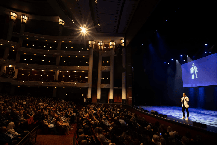 On stage, Matt Fraser addresses a large audience in a dimly lit theater. The upper balconies are visible, and a spotlight highlights him as he speaks. A screen displays Matt Fraser's image.