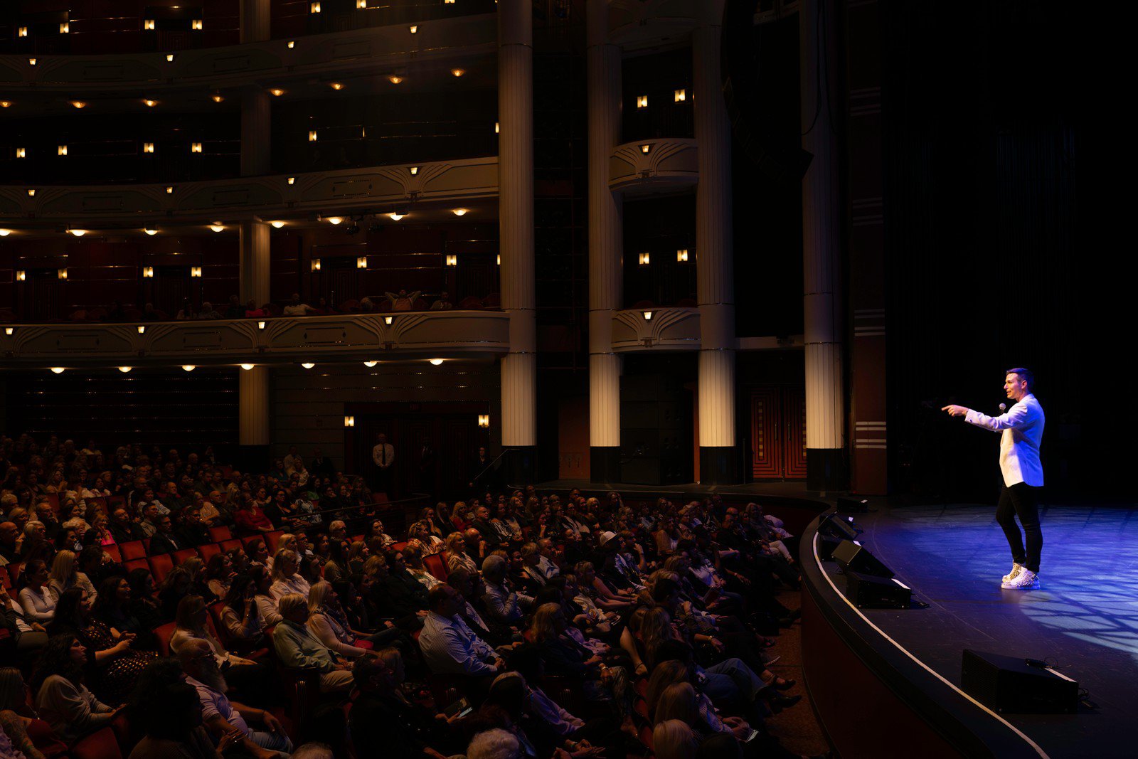 Matt Fraser addresses a large seated audience in a theater with balcony seating. The stage is brightly lit while the audience is dimly lit.