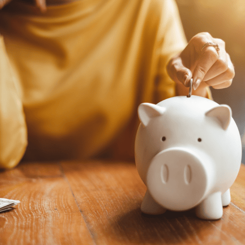 A person wearing a yellow shirt, inserts a coin into a white piggy bank on a wooden surface.