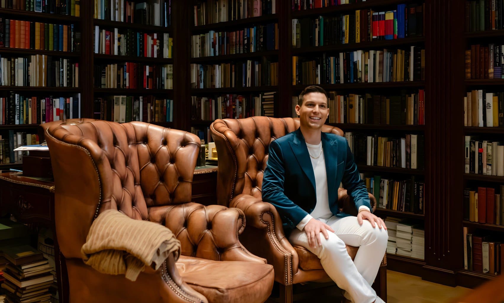Psychic Medium Matt Fraser sits on a brown leather chair in a library with shelves full of books. Another identical chair is empty beside him. He is smiling and wearing a dark jacket and light-colored pants.