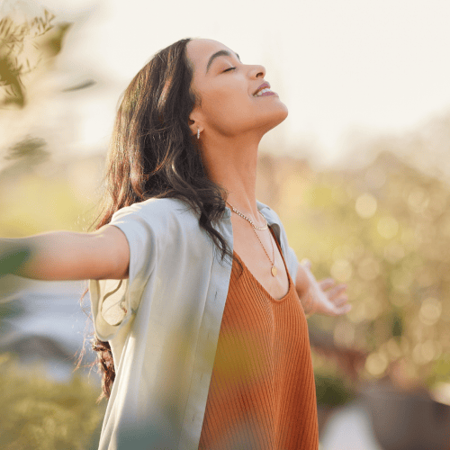 A woman with long hair stands outdoors with arms outstretched and eyes closed, appearing relaxed. Matt Fraser is visible alongside her, wearing a light shirt over an orange top. The background is blurred and sunny.