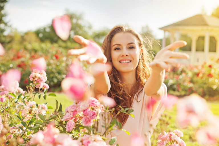 A woman joyfully extends her arms amidst blooming pink flowers in a sunny garden, with Matt Fraser in the background near a gazebo.