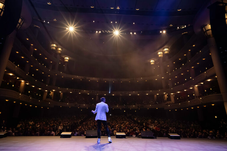 Matt Fraser stands on stage in a theater, facing a large audience under bright lights.