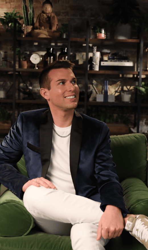 Matt Fraser, in a blue blazer and white pants, sits on a green couch, smiling, with shelves filled with plants and decorative items in the background.
