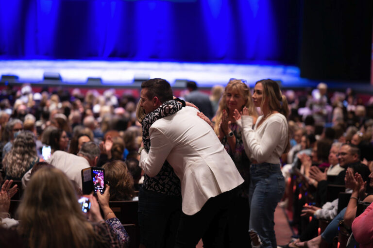 Matt Fraser, wearing a white jacket, hugs another person in a crowded auditorium. Audience members are seated, some are standing, and a few are capturing the moment on their phones.