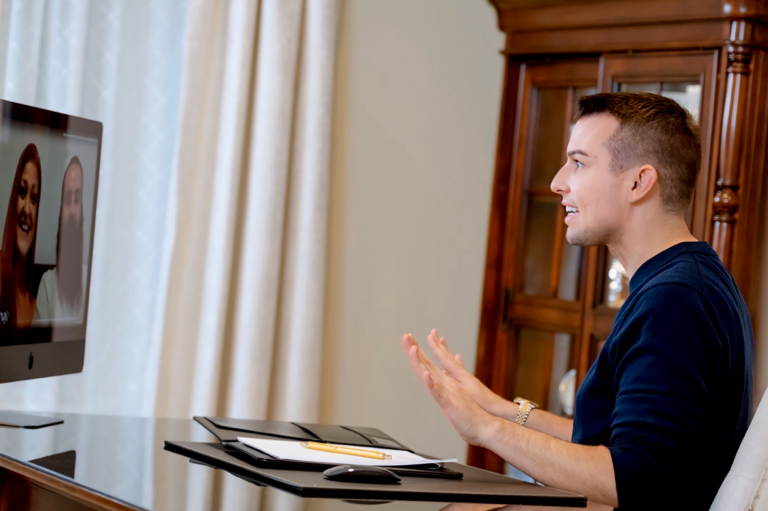 Matt Fraser, wearing a blue sweater, talks via video call on a computer. A notebook and pen are on the desk, and two people appear on the screen.