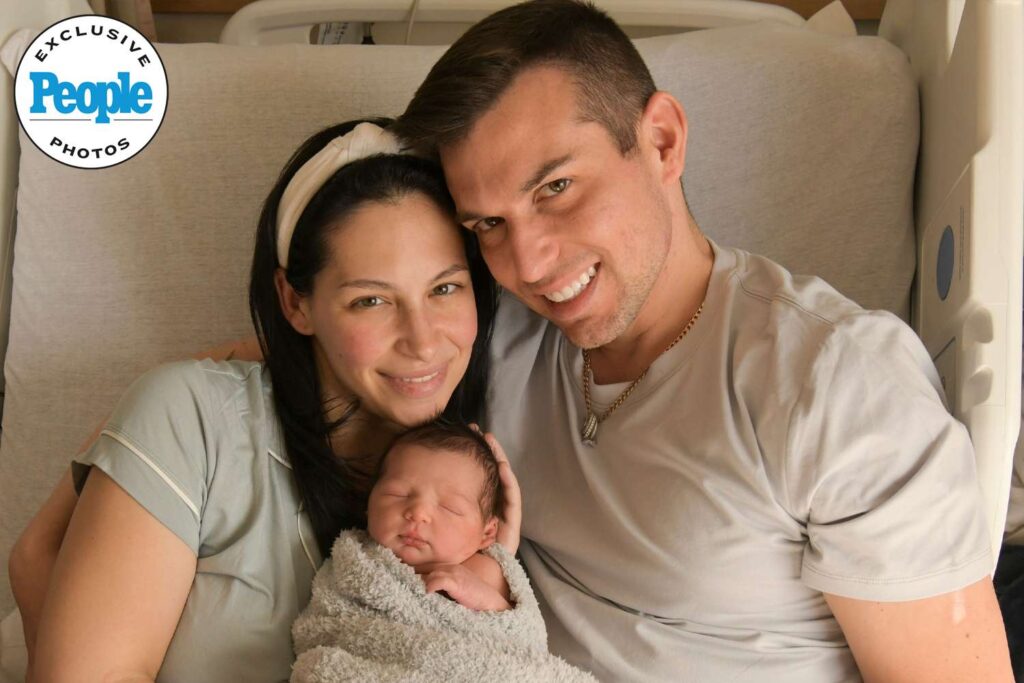 Sitting on a hospital bed, with Matt Fraser holding a sleeping newborn wrapped in a gray blanket.