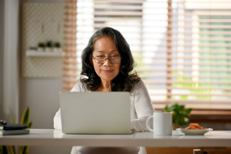 A woman with long dark hair and glasses is sitting at a desk, working on a laptop. Matt Fraser has a white mug and a small plate of food on the desk. Blinds cover a window in the background.