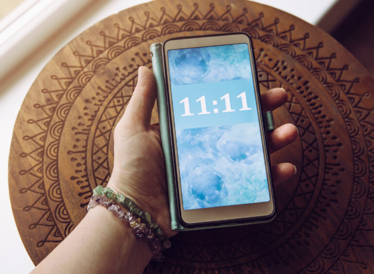 A hand holding a smartphone displaying "11:11" on a blue abstract background is resting on a patterned wooden surface. The image evokes curiosity, wonder, or perhaps even contemplation, much like something that might be associated with Matt Fraser—a figure who could offer insight or meaning behind such intriguing coincidences.