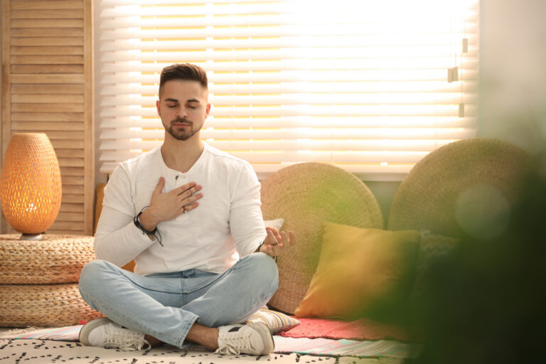 man dressed in a white shirt and jeans, sits cross-legged on a rug. He is meditating with one hand on his chest in a sunlit room adorned with woven decor and blinds.