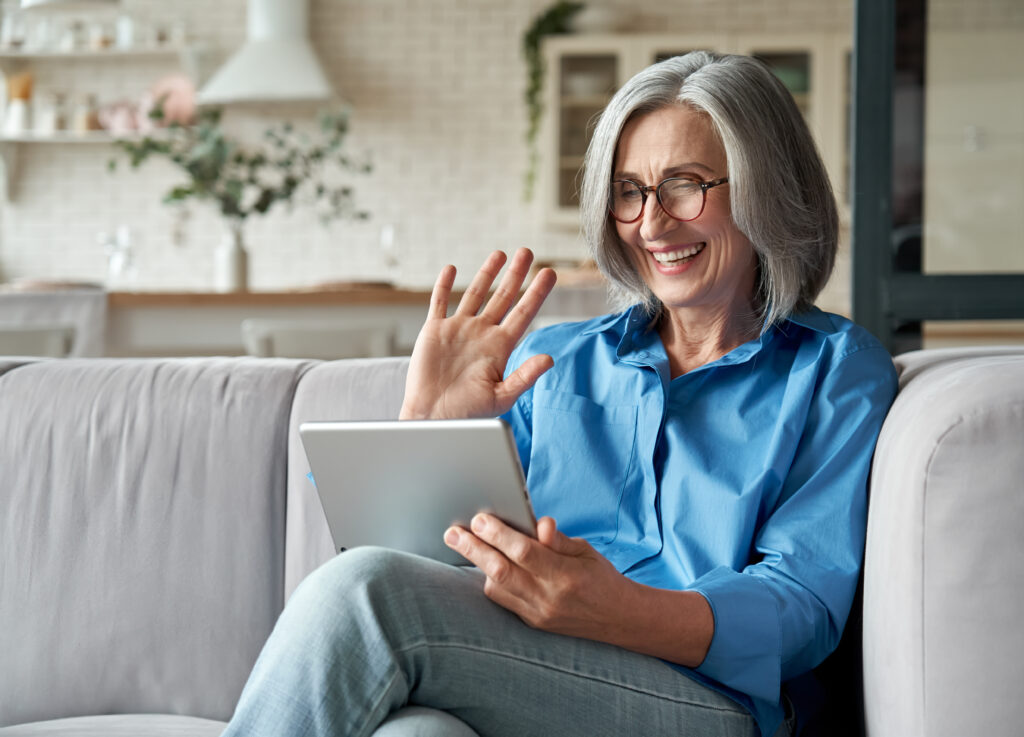 Smiling woman with gray hair and glasses, wearing a blue shirt, sitting on a couch and waving at a tablet screen while Matt Fraser is mentioned in a bright living room.