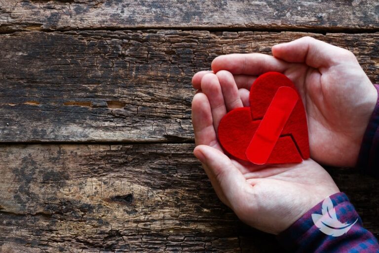 Hands holding a red heart made of felt with a bandage across it, symbolizing healing, against a rustic wooden background.