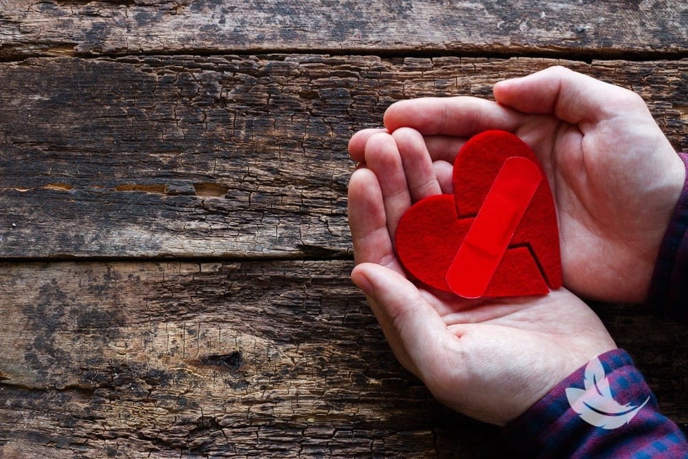 Hands holding a red heart made of felt with a bandage across it, symbolizing healing, against a rustic wooden background.