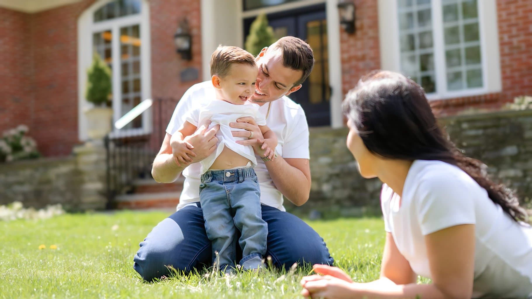 Matt Fraser and Alexa Fraser play with Royce Fraser on a grassy lawn in front of a brick house. Matt Fraser is tickling the boy, and the woman is watching them.